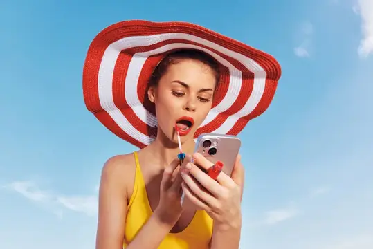 Молодая женщина в жёлтом купальнике наносит помаду, держа в руках смартфон, под голубым небом