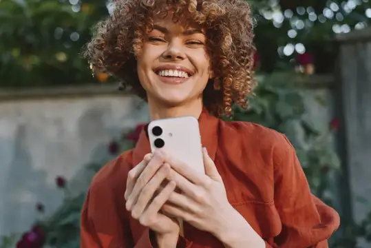 Улыбающаяся кудрявая девушка держит смартфон в саду