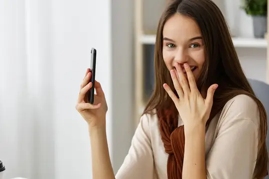 Молодая женщина смеется, закрывая рот рукой, глядя на смартфон