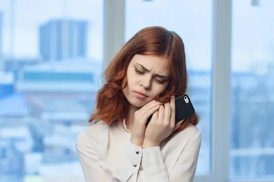 Рыжеволосая женщина с закрытыми глазами слушает телефон у окна
