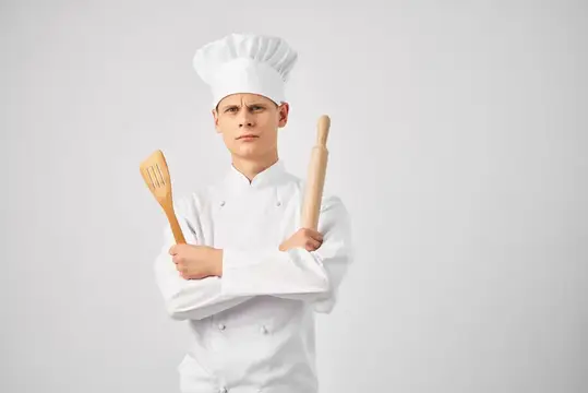 Мужчина-повар в колпаке на голове на фоне кухонной утвари и светлого фона