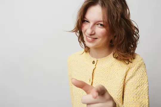 Счастливая женщина с вьющимися волосами в желтом вязаном свитере указывает прямо в камеру с улыбкой