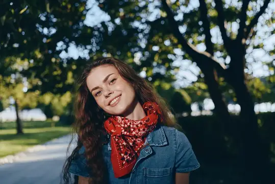 Улыбающаяся женщина в красном шарфе стоит в залитом солнцем парке, одетая в повседневную синюю одежду, излучая естественную радость и расслабленное настроение среди зеленых деревьев
