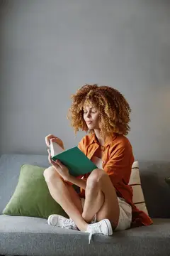 Кудрявая девушка в оранжевой рубашке читает книгу на диване