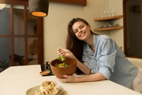 Улыбающаяся темноволосая девушка ест салат из глиняной миски на светлой кухне