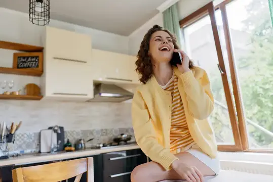 Кудрявая девушка в жёлтом кардигане весело смеётся, разговаривая по телефону на светлой кухне