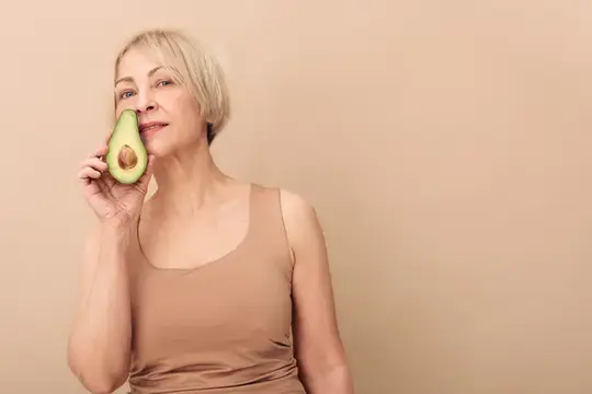 Светловолосая женщина с авокадо у лица на бежевом фоне