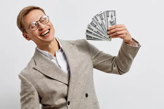 Улыбающийся счастливый светлокожий мужчина в очках и сером пиджаке держит веер денежных купюр с радостным выражением лица на однотонном белом студийном фоне