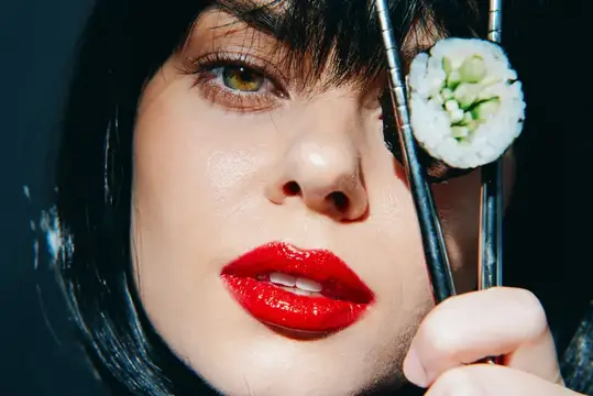 Крупный план портрета молодой женщины с ярко-красными губами, держащей суши палочками на темном фоне, вызывающий ассоциации с современной кулинарной культурой и элегантностью