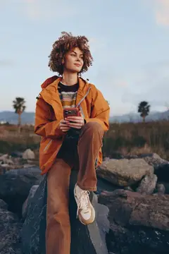 Кудрявая девушка в оранжевой куртке сидит на камнях с телефоном
