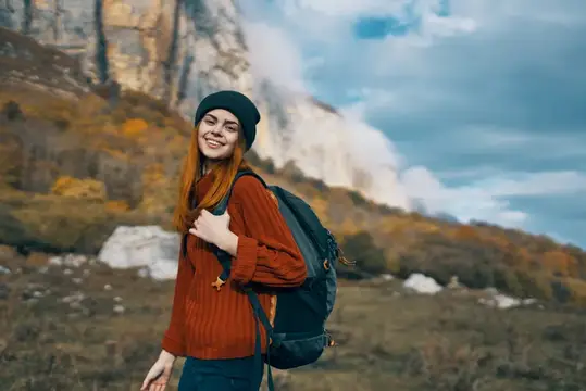 Рыжеволосая девушка с рюкзаком улыбается на фоне осенних гор