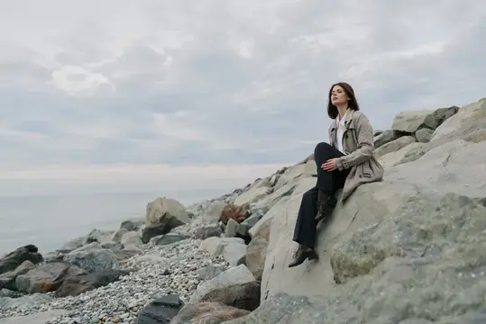 Безмятежная женщина в куртке сидит на каменистом берегу у океана