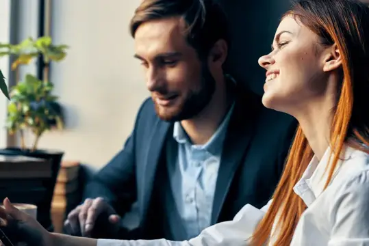 Улыбающиеся мужчина и рыжеволосая женщина работают за ноутбуком в офисе