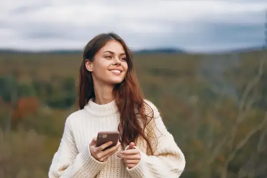 Улыбающаяся девушка в белом свитере с телефоном на осенней прогулке