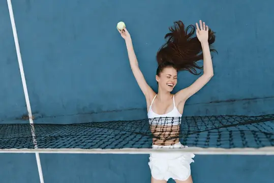 Счастливая молодая женщина в спортивной одежде наслаждается игрой в теннис, демонстрируя свое волнение с радостным выражением лица на фоне ярко-синего фона, который передает динамичную летнюю…
