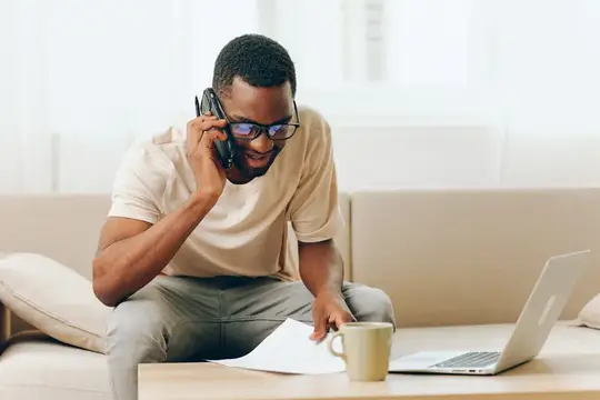 Улыбающийся афроамериканец работает за ноутбуком, сидя на диване в своей современной квартире