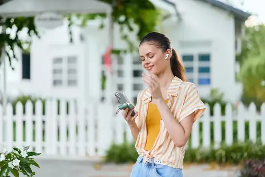 Улыбающаяся девушка в наушниках смотрит в смартфон у белого забора