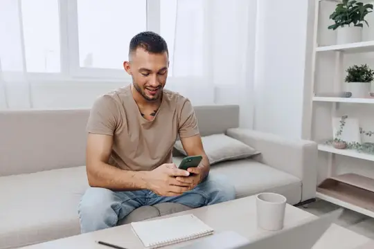 Молодой мужчина улыбается, глядя в смартфон на диване