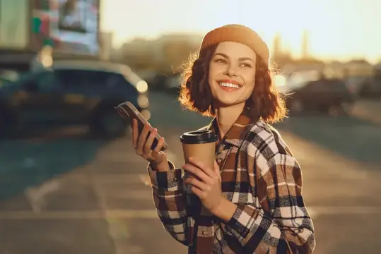 Улыбающаяся девушка в берете с кофе и телефоном на закате