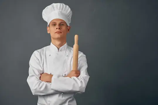 Молодой повар в белом колпаке с деревянной скалкой на сером фоне