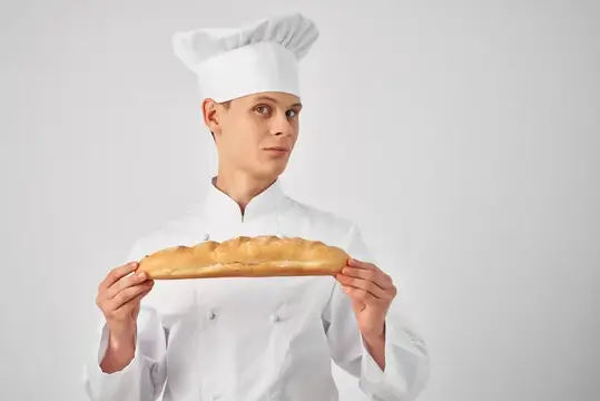 Повар в белом колпаке держит длинный багет на сером фоне