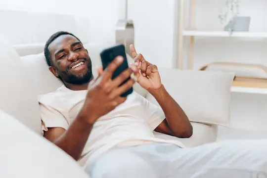 Улыбающийся темнокожий мужчина лежит на диване со смартфоном
