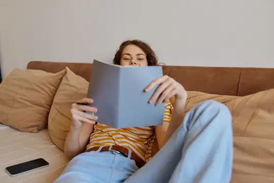 Молодая женщина читает книгу, лёжа на диване с подушками