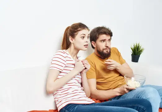 Забавные мужчина и женщина отдыхают дома на диване, веселятся с попкорном