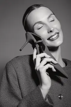 Улыбающаяся женщина европеоидной расы со светлой кожей и закрытыми глазами нежно держит туфлю на высоком каблуке у лица, на ней фактурный свитер, в студии