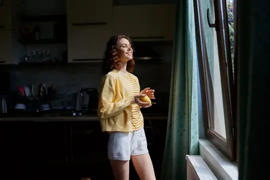 Улыбающаяся девушка с яблоком стоит у окна на кухне