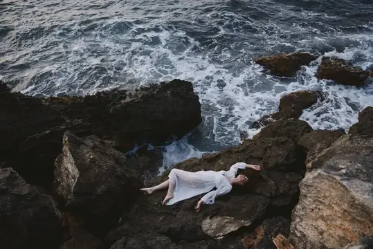 Женщина в белом платье лежит на камнях у бурного моря