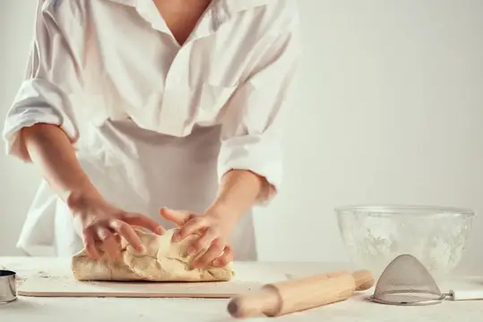 Повар в белом фартуке месит тесто на кухонном столе