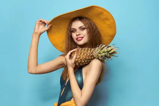 Женщина в пляжной шляпе держит ананас и купальник на фоне экзотических фруктов и голубого фона