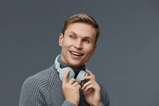 Улыбающийся светловолосый парень в свитере с наушниками на шее
