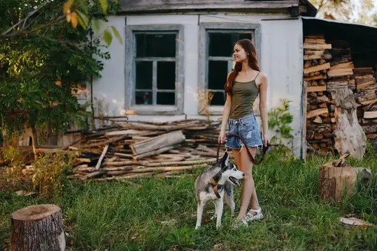 Женщина и её хаски весело играют на улице в парке среди деревьев, улыбаясь, показывая зубы, во время осенней прогулки со своим питомцем