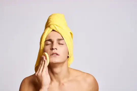 Красивый мужчина с косметологической губкой для лица и желтым полотенцем на голове
