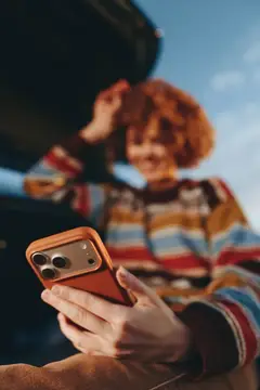 Руки держат смартфон в оранжевом чехле на фоне размытой рыжеволосой девушки