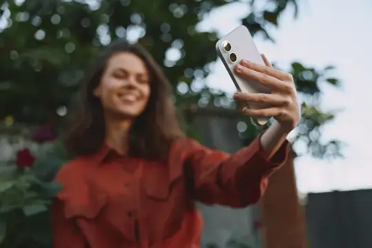 Улыбающаяся девушка в красной рубашке делает селфи на смартфон в саду