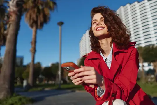 Счастливая молодая женщина в красной куртке пользуется смартфоном на улице в солнечный день; за ней пальмы и городские здания, она наслаждается свежим воздухом и технологиями