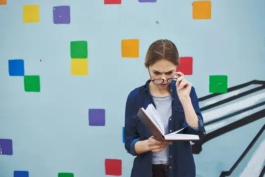 Девушка в очках читает книгу на фоне стены с цветными квадратами