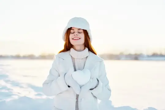 Рыжеволосая девушка в белой шапке держит снежное сердце зимой