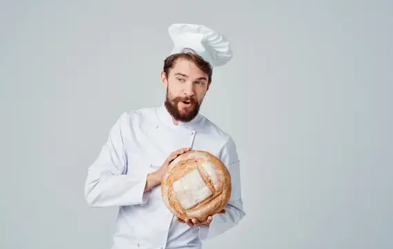 Шеф-повар в светлом костюме и головном уборе держит в руке круглый каравай хлеба