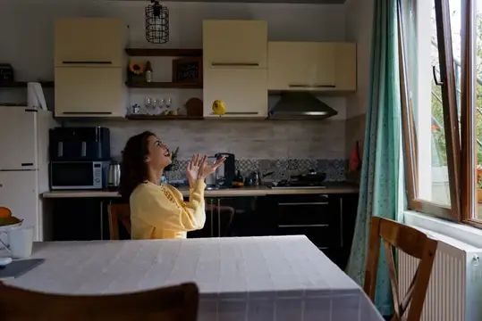 Жизненная домашняя сцена с улыбающейся женщиной в светлой кухне, непринужденная поза, естественный свет, мягкие цвета, повседневный момент, расслабленная атмосфера, чистые столешницы, открытое окно