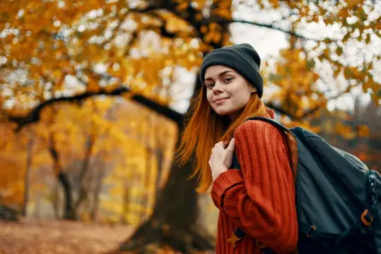 Рыжеволосая девушка в шапке с рюкзаком на фоне осенних деревьев