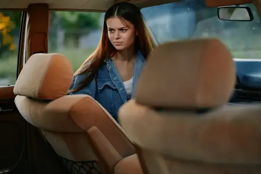 Задумчивая молодая женщина в джинсовой куртке сидит в старом автомобиле, запечатлевая момент размышлений в мягком свете и винтажной атмосфере