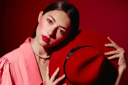 Креативный beauty-портрет стильной молодой женщины, держащей красную широкополую шляпу, в розовой блузе, с яркими глянцевыми губами, жемчужным ожерельем и минимальным макияжем на ярко-красном фоне