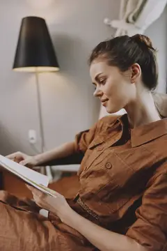 Молодая женщина в коричневом платье читает книгу в кресле у торшера