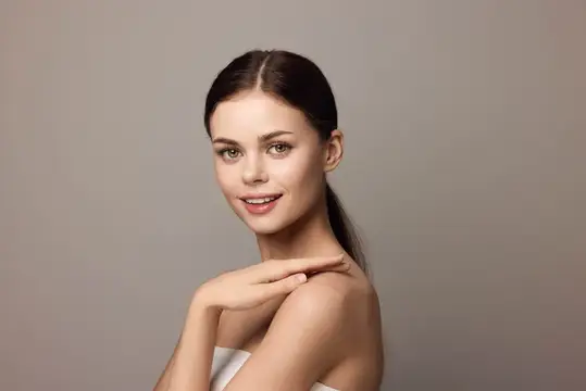 Молодая темноволосая женщина с улыбкой касается плеча на сером фоне