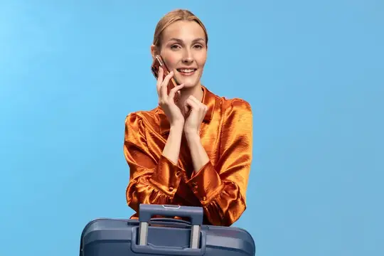 Улыбающаяся блондинка в оранжевой блузе говорит по телефону с чемоданом