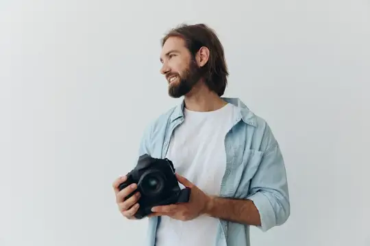 Мужчина-хипстер-фотограф в студии на фоне белого фона держит профессиональную камеру и настраивает её перед съёмкой